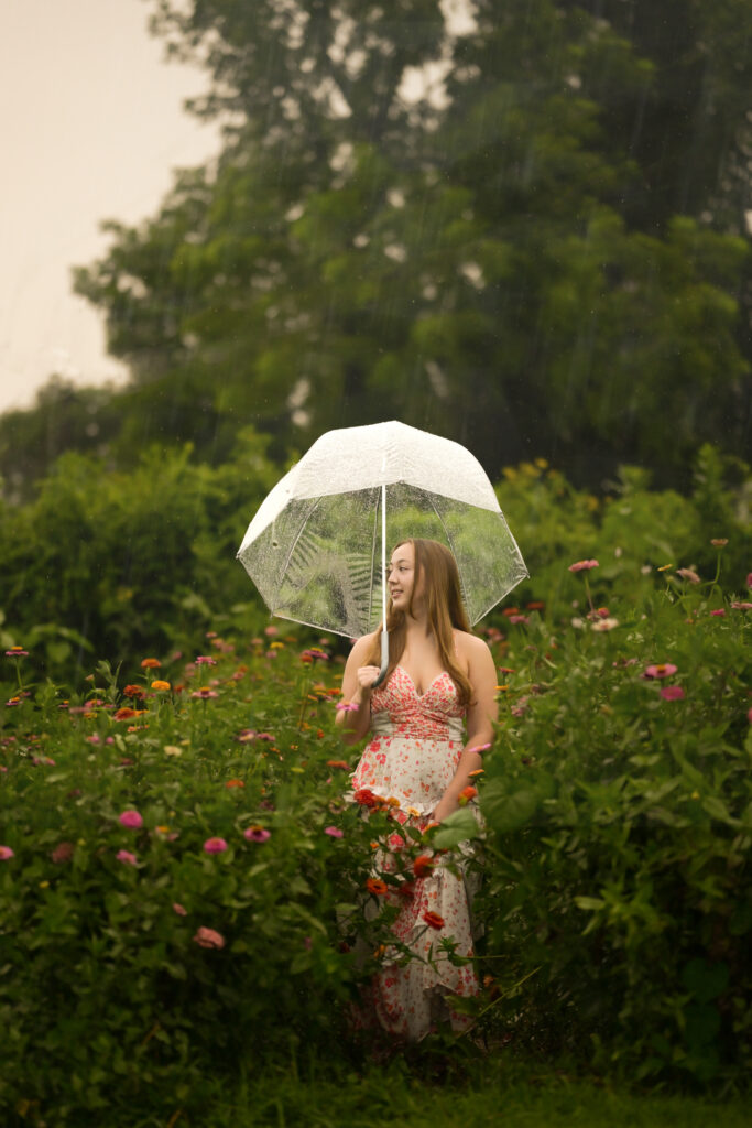 Young woman in floral dress holding clear umbrella in colorful flower garden on rainy day.